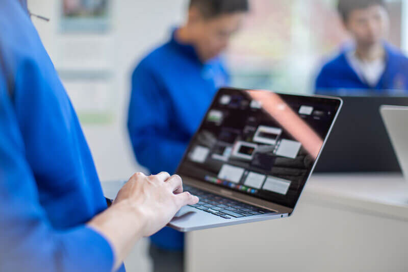 Person using a laptop, with two people blurred in the background in a modern, bright setting.