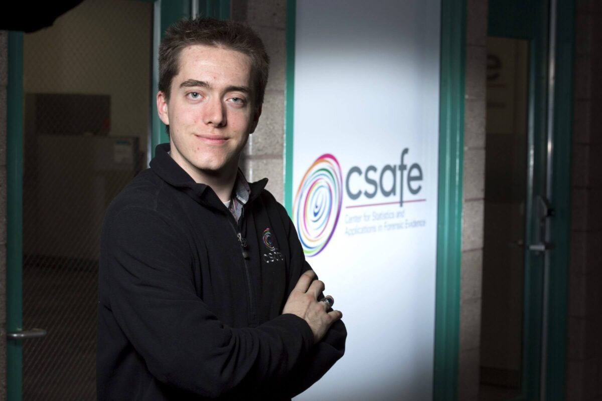 A person stands with folded arms, smiling in front of a sign that reads "csafe.