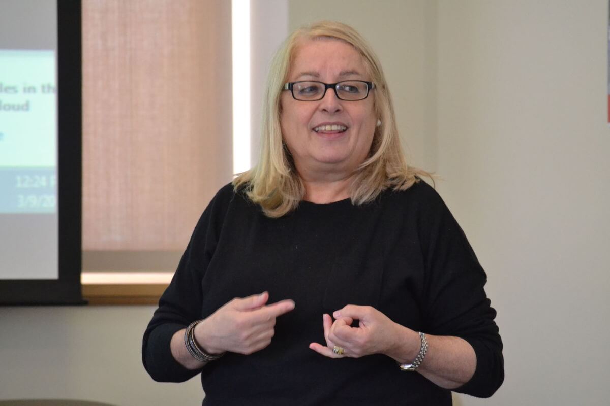 A woman with blonde hair and glasses gestures while speaking in a classroom or meeting room.