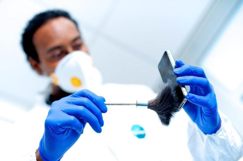Person in gloves and mask dusting a smartphone for fingerprints in a forensic investigation lab.