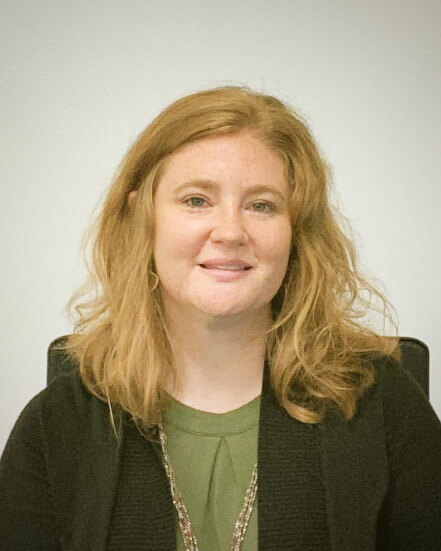 A woman with wavy red hair, wearing a green top and black cardigan, sits and smiles at the camera.