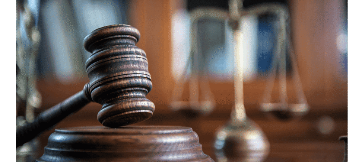 A wooden judge’s gavel rests on a sound block, with scales of justice blurred in the background.