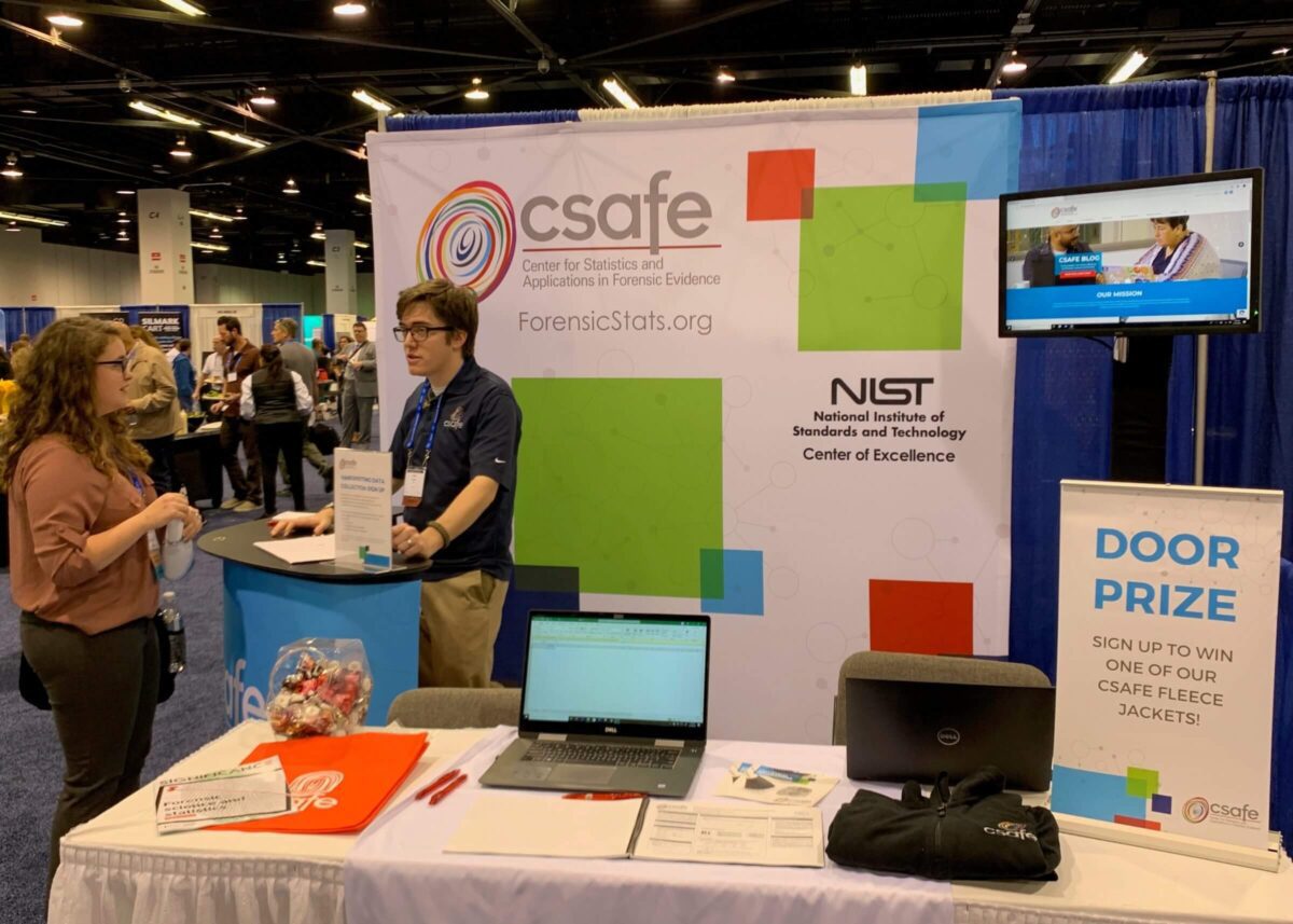Two people talk at a CSAFE expo booth with displays, brochures, and a door prize sign.