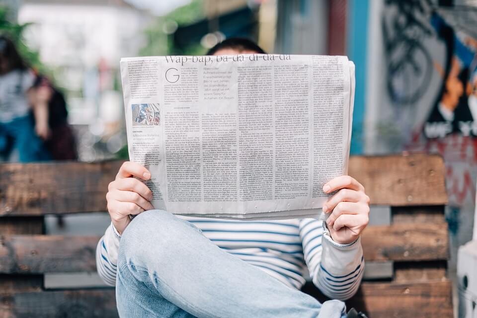 Person sitting on a bench, reading a newspaper, face hidden behind the paper.