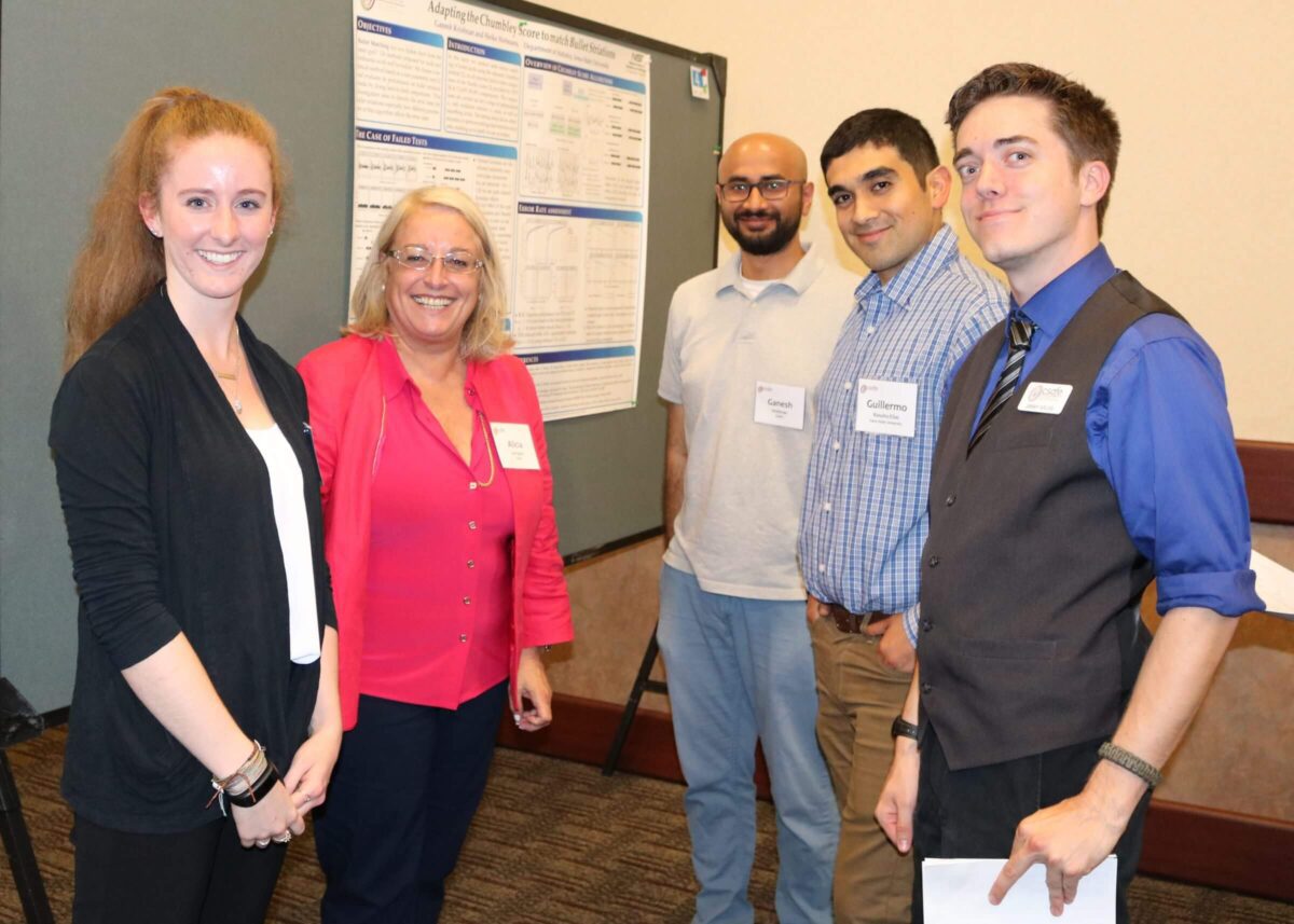 Five people stand and smile in front of a scientific poster at a presentation or conference event.