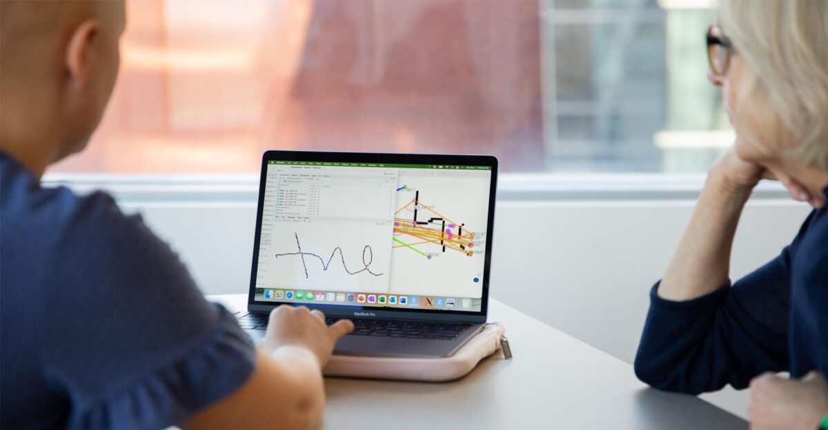 Two people look at a laptop screen displaying code, a sketch, and a 3D structure in a bright room.