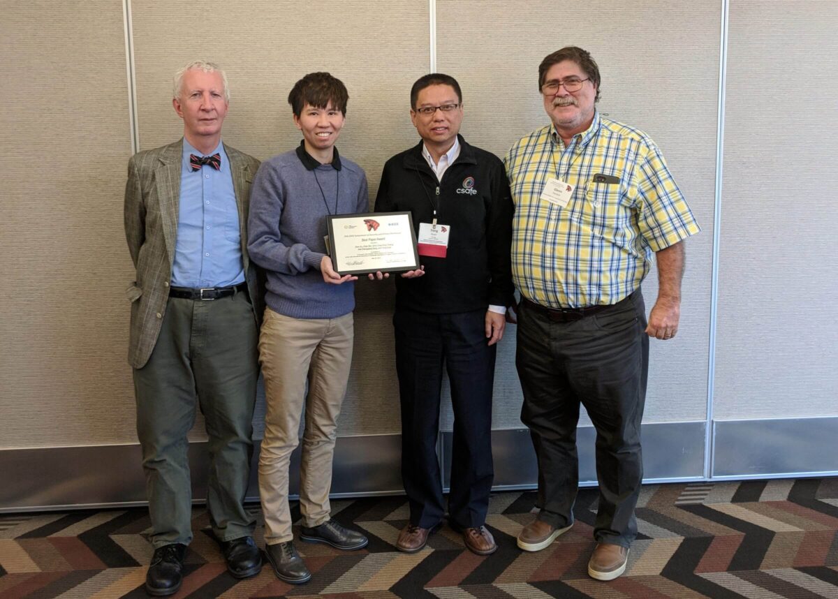 Four people stand indoors; one holds a plaque. They are posing and smiling for a group photo.