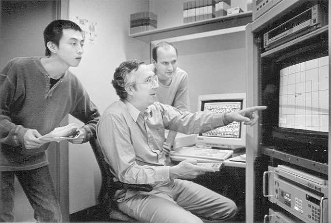 Three men analyze data on monitors and equipment in a computer lab or control room.