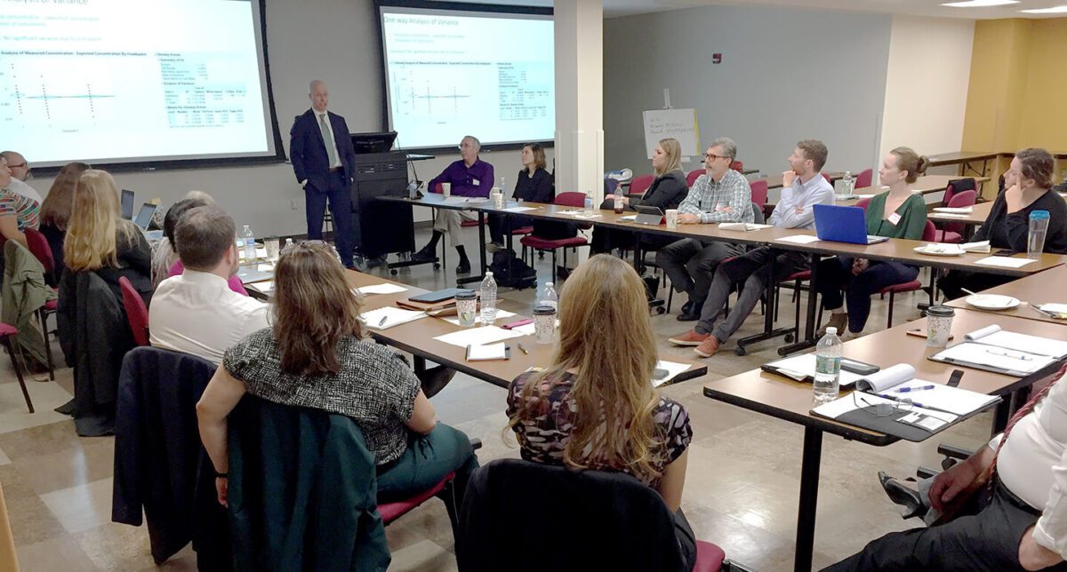 A man presents data on a screen to a group of people seated around tables in a meeting room.