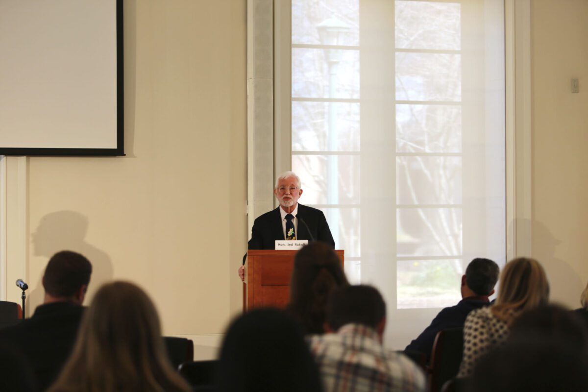 An older man speaks at a podium to an audience in a bright room with large windows behind him.