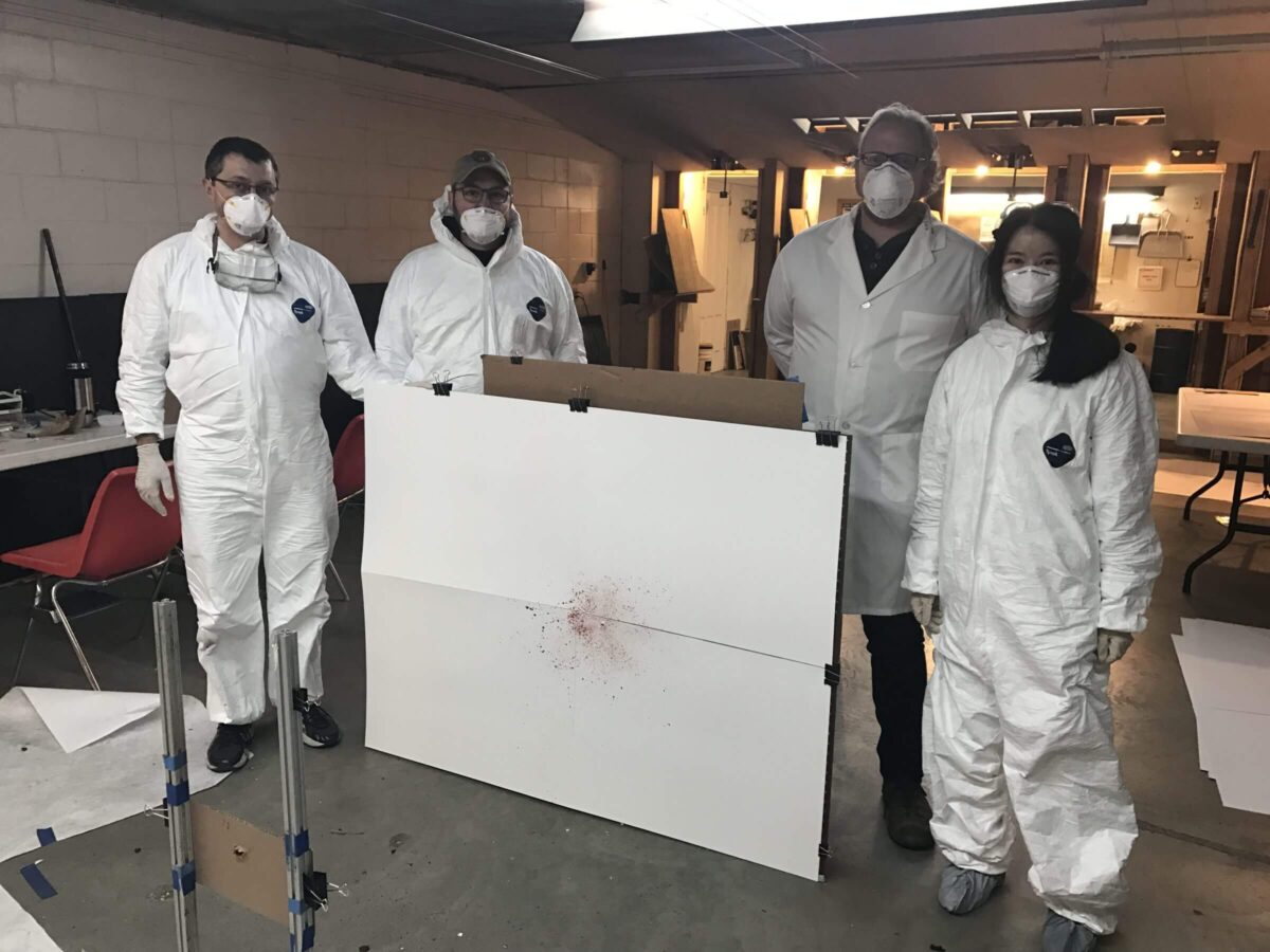 Four people in protective suits stand around a white board with a red splatter in a lab.