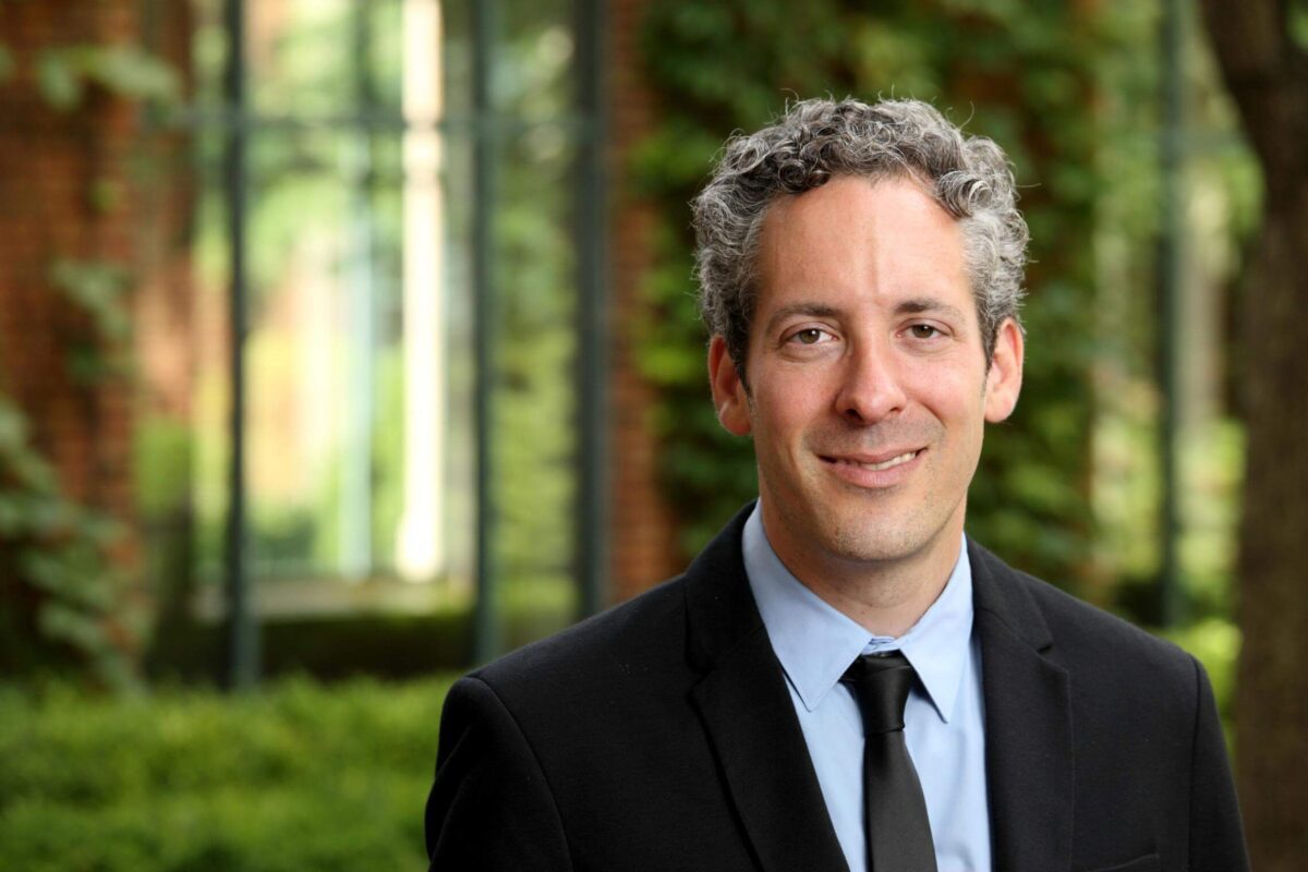 Smiling man in a black suit and tie stands outdoors with greenery and brick walls in the background.