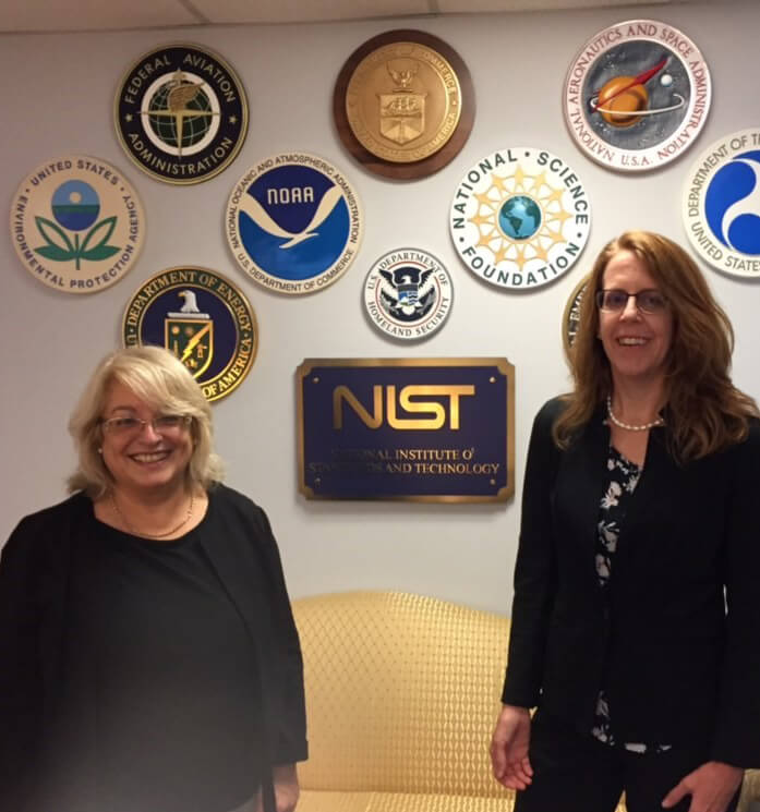 Two women standing in front of a NIST sign and various U.S. government agency logos on a wall.