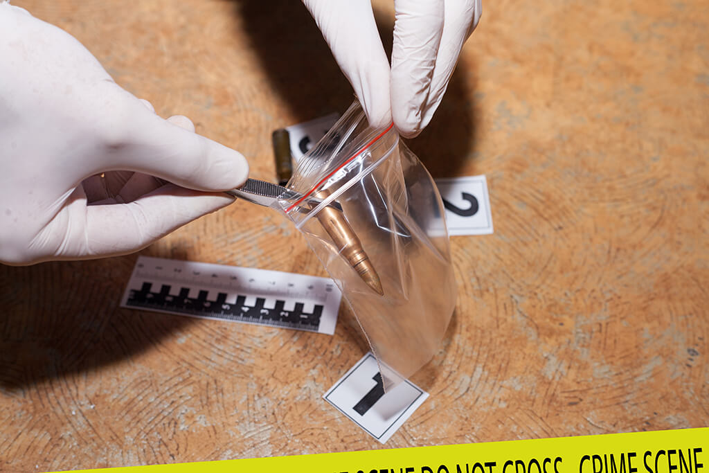 Gloved hands bag a bullet as evidence at a crime scene with numbered markers and a ruler on the floor.