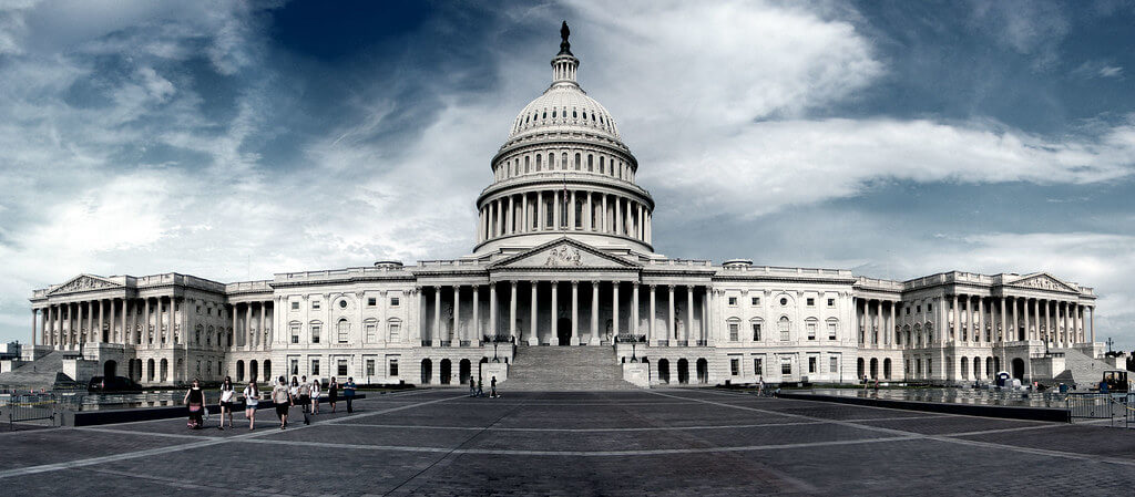 The United States Capitol building with a few people walking in front under a cloudy sky.
