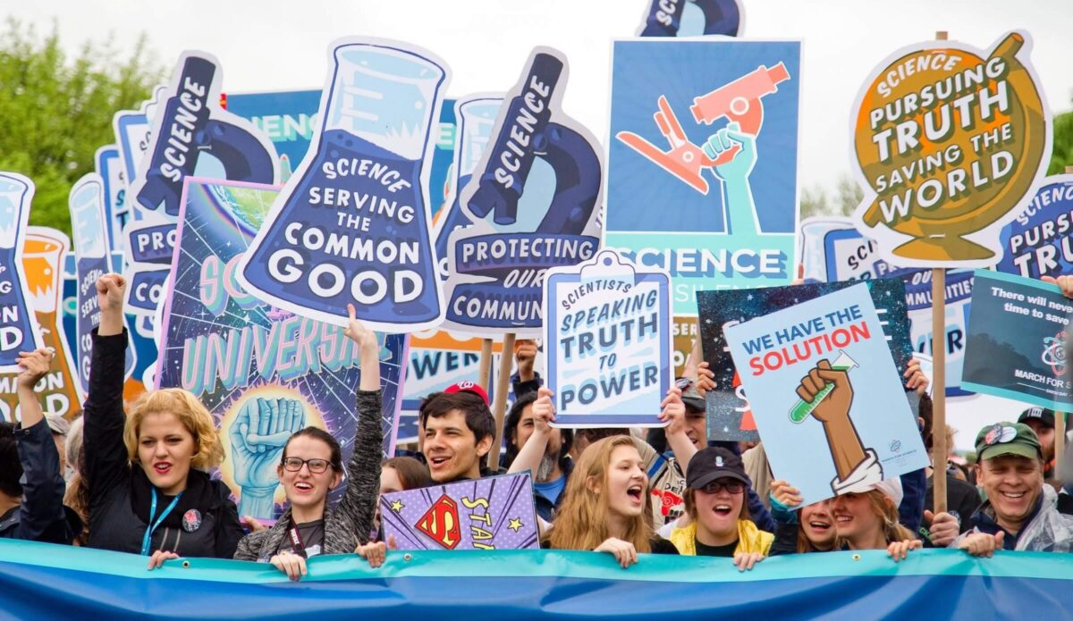 A diverse crowd holds science-themed signs at a rally, advocating for truth and the common good.