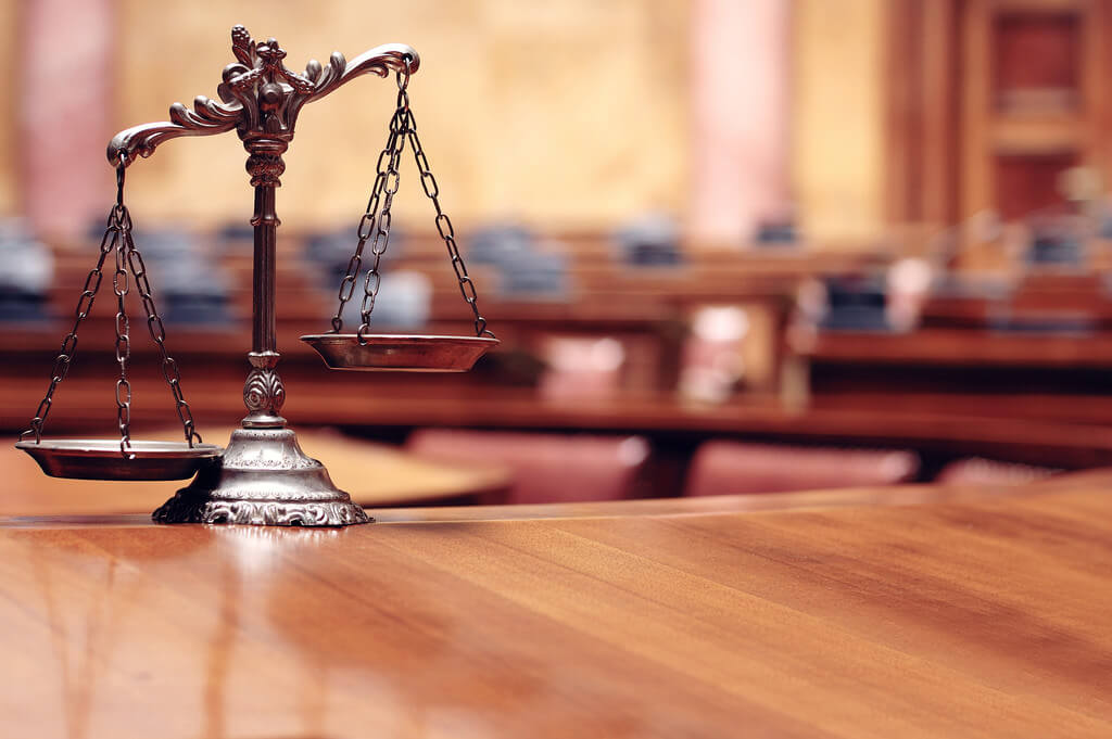 A bronze balance scale sits on a wooden desk in an empty courtroom.