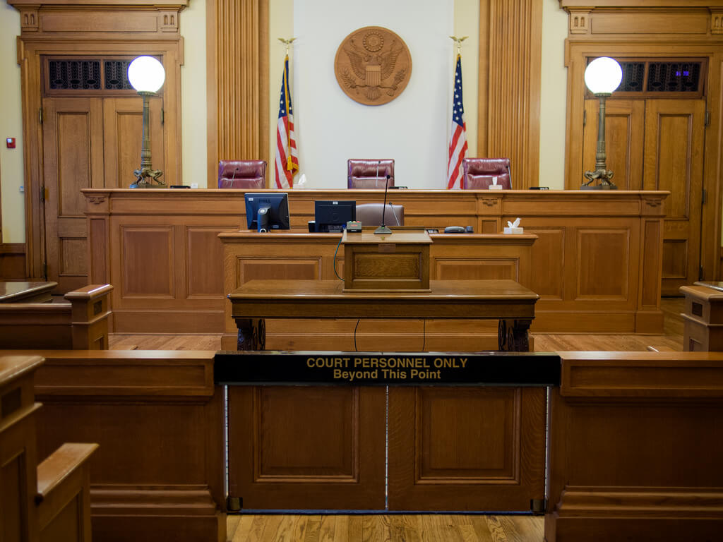Empty courtroom with wooden benches, judge's desk, and two American flags behind the bench.