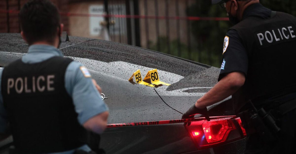 Two police officers examine a car marked with evidence tags at a crime scene on a rainy day.
