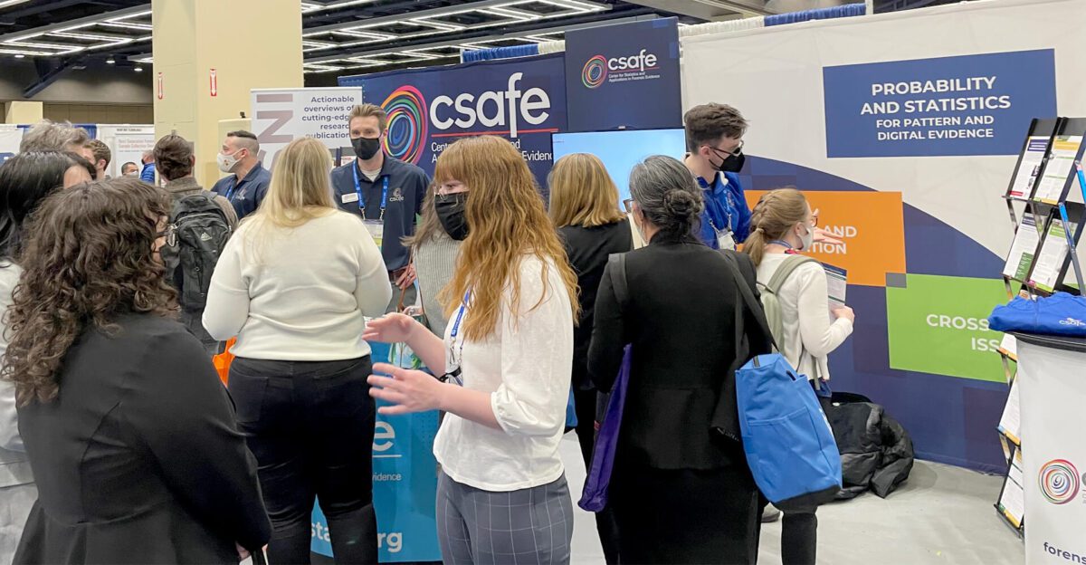 People gather and talk at a conference booth with "csafe" and "Probability and Statistics" banners.