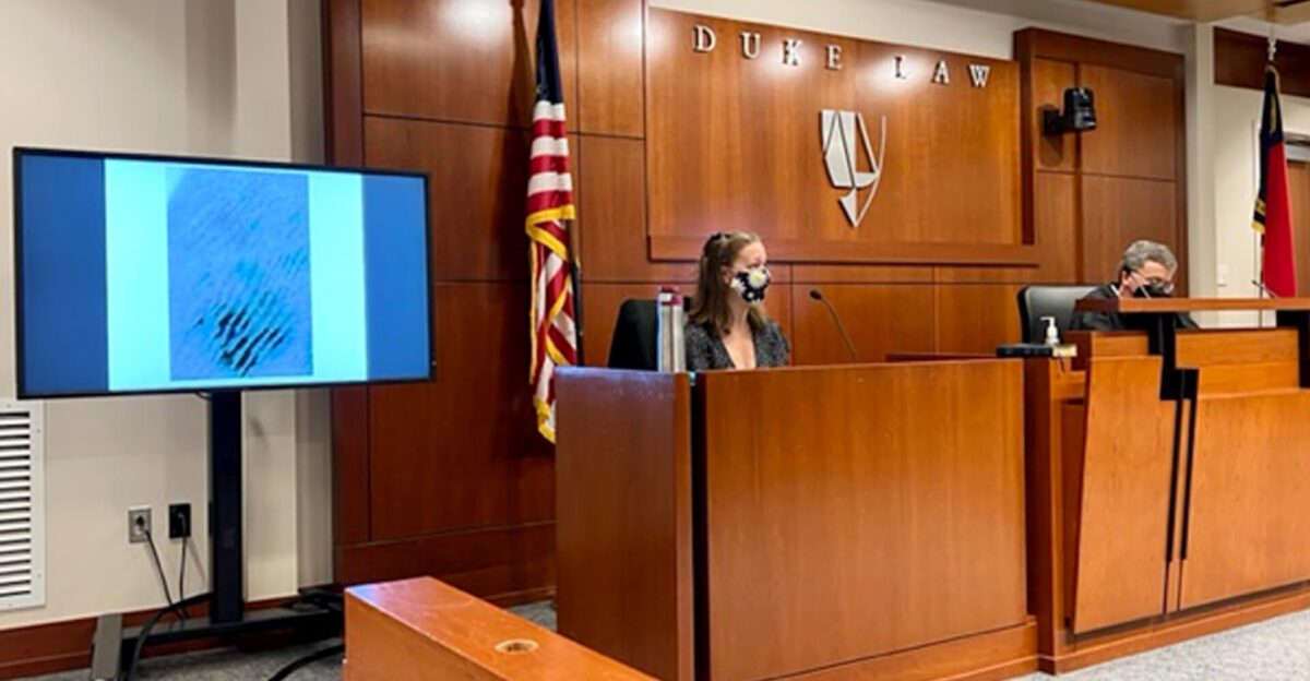 A woman testifies in a courtroom at Duke Law as an image is displayed on a screen beside her.