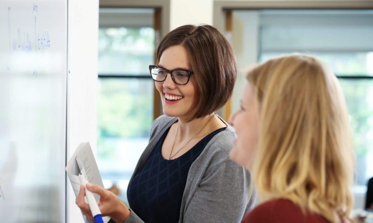 Two women smiling and collaborating at a whiteboard in a bright, modern office setting.