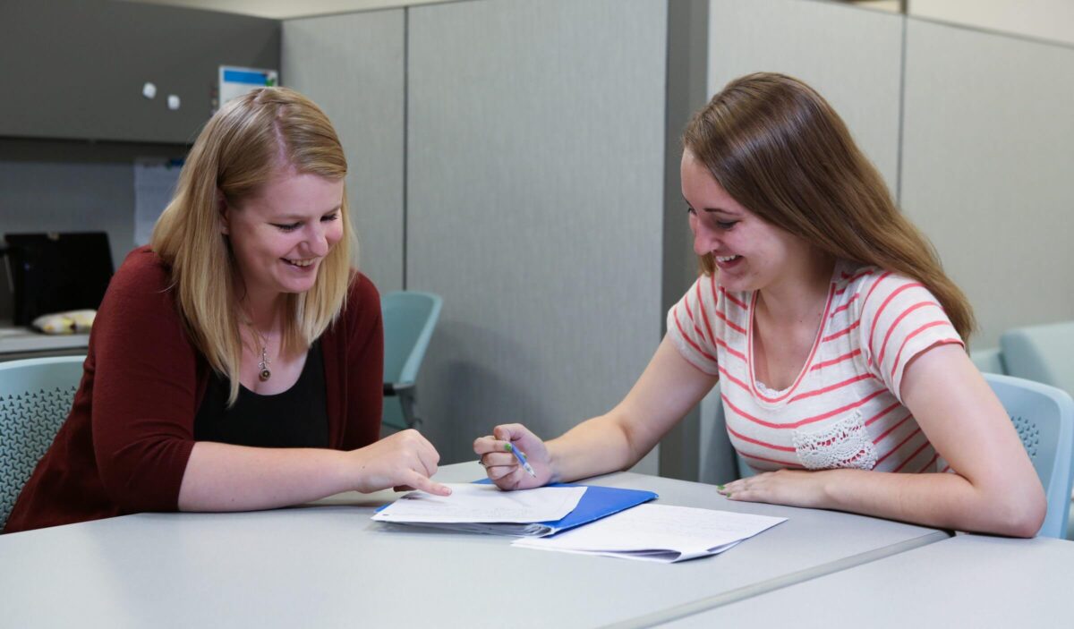 Two women sit at a table smiling and reviewing documents together in an office setting.