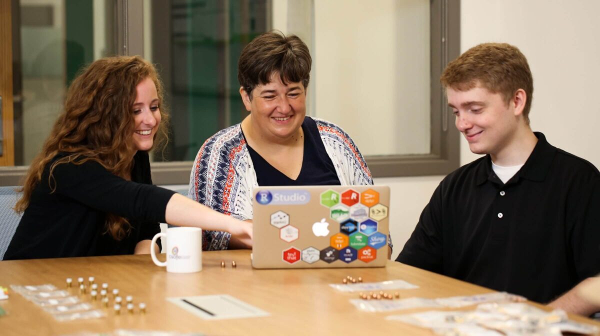 Three people smiling and working together at a table with a laptop, small parts, and a coffee mug.