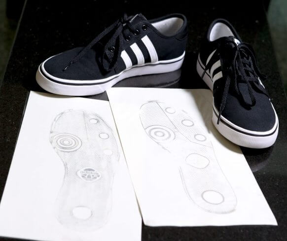 Two black sneakers with white stripes placed next to paper sketches of their outsoles on a dark surface.