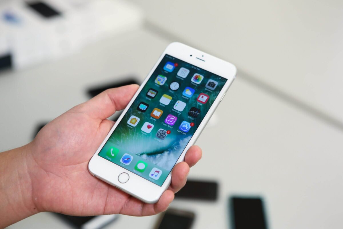 A hand holding a white smartphone displaying various app icons on its home screen.