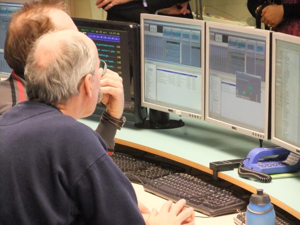 Two men sit at desks, looking at multiple computer monitors displaying data and charts.