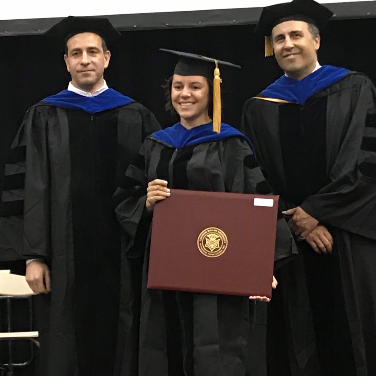 Three people in graduation robes, one holding a diploma and smiling, standing indoors at a ceremony.