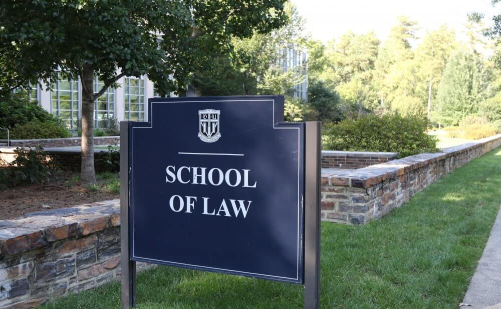 A blue sign reading "School of Law" stands on a grassy area near a building and trees.