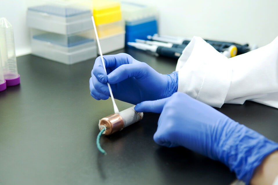 Gloved hands using a cotton swab to collect a sample from a surface in a laboratory setting.
