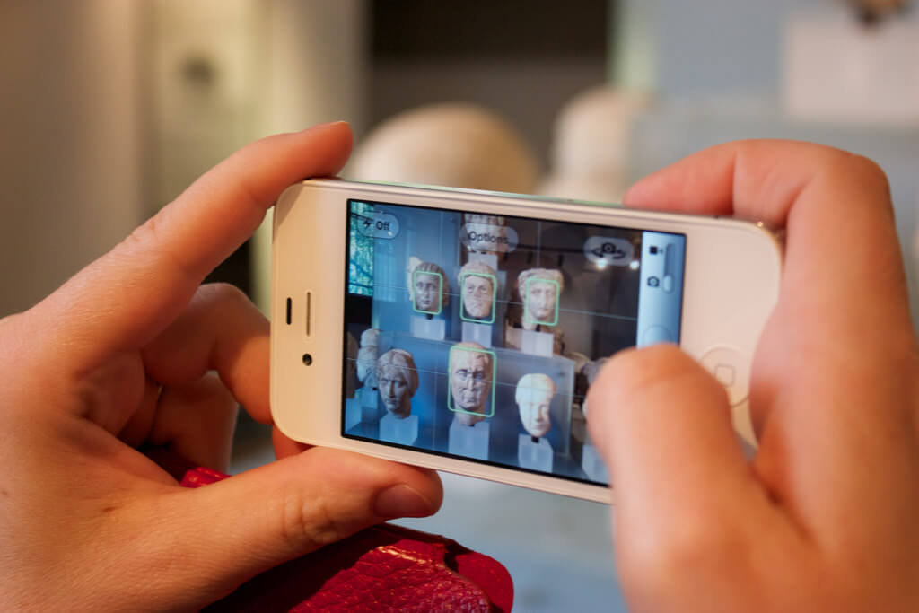 Hands holding a smartphone, displaying an app that shows ancient busts with overlaid digital labels.
