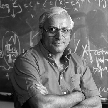 An older man with glasses stands in front of a chalkboard covered in mathematical equations.