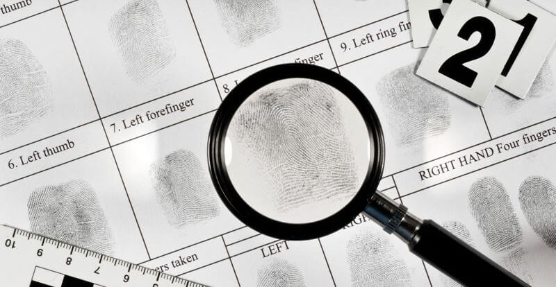 A magnifying glass over fingerprint sheets with evidence markers and a ruler nearby.