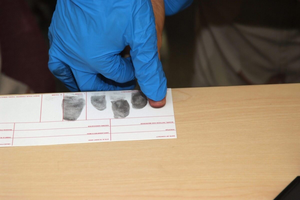 A gloved hand is taking fingerprints on a white fingerprint card on a light wooden surface.