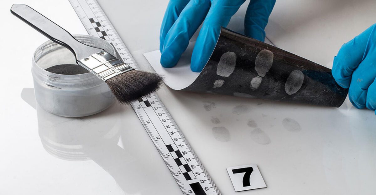 Gloved hands lift film with fingerprints, brush, powder, ruler, and evidence marker visible.