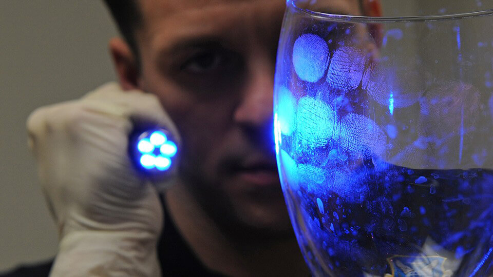A gloved person shines a blue light on a glass revealing visible fingerprints on its surface.
