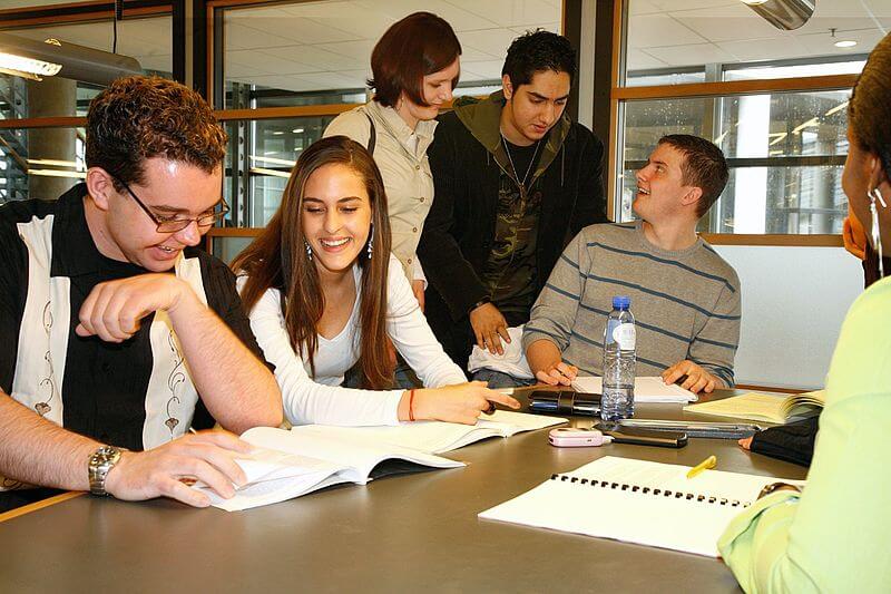 Six students smiling and studying together at a table with books and notebooks in a classroom.