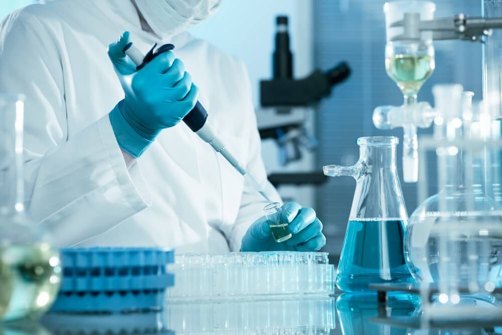 Scientist in lab coat and gloves using pipette with test tubes and beakers on laboratory table.