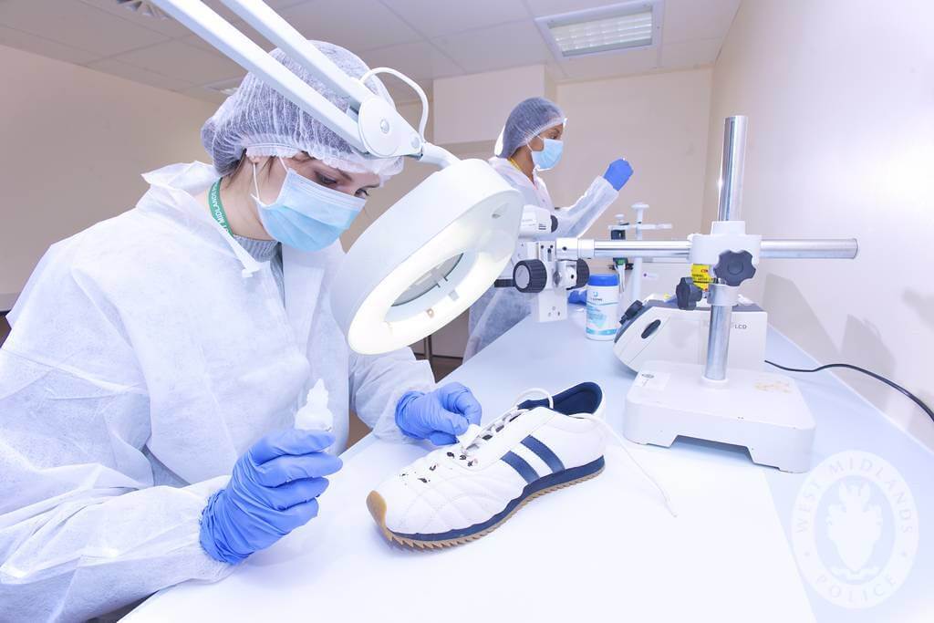 Two forensic scientists examine a white sneaker in a lab, wearing protective clothing and gloves.