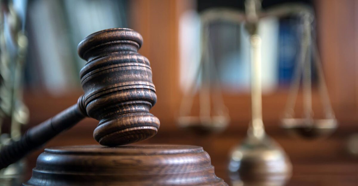 A wooden judge’s gavel rests on a sound block with blurred scales of justice in the background.
