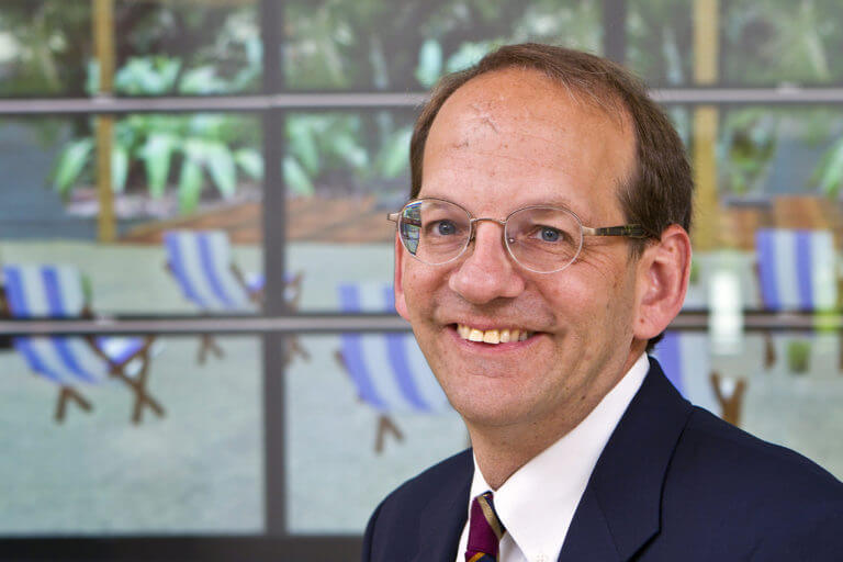 Smiling man in glasses and suit, with blurred beach chairs and greenery in the background.