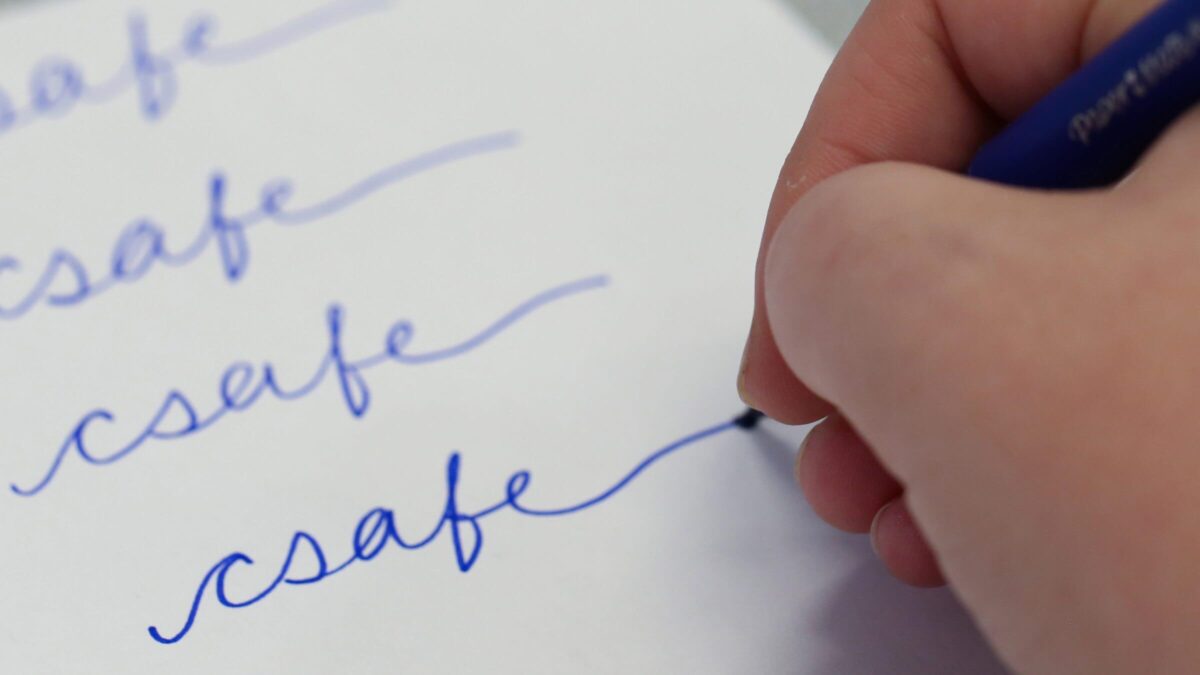 A hand writing the word "csafe" repeatedly in blue ink on white paper.