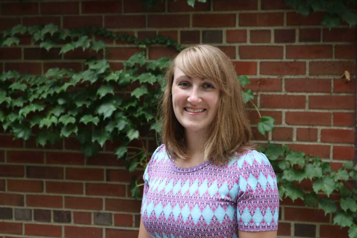 Smiling woman with shoulder-length hair stands in front of a brick wall with green ivy leaves.