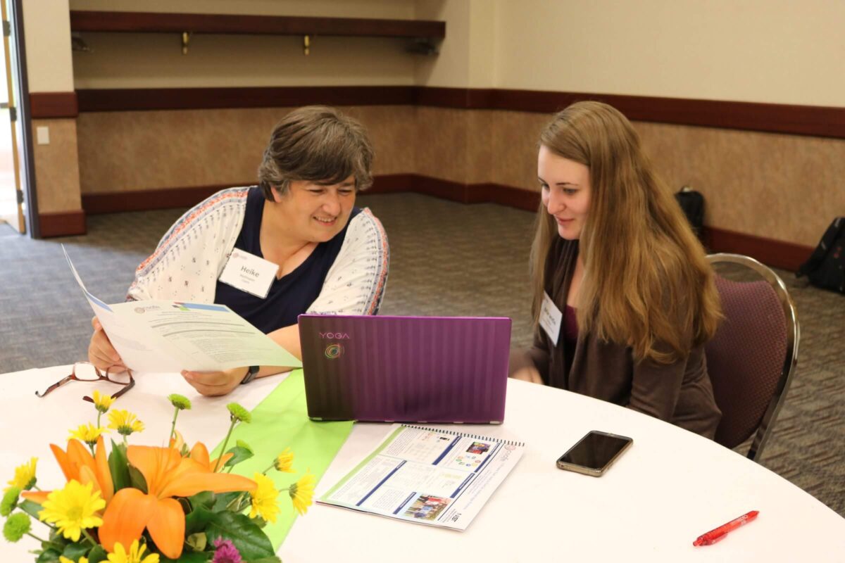 Two women sit at a round table looking at a purple laptop, with papers and flowers on the table.