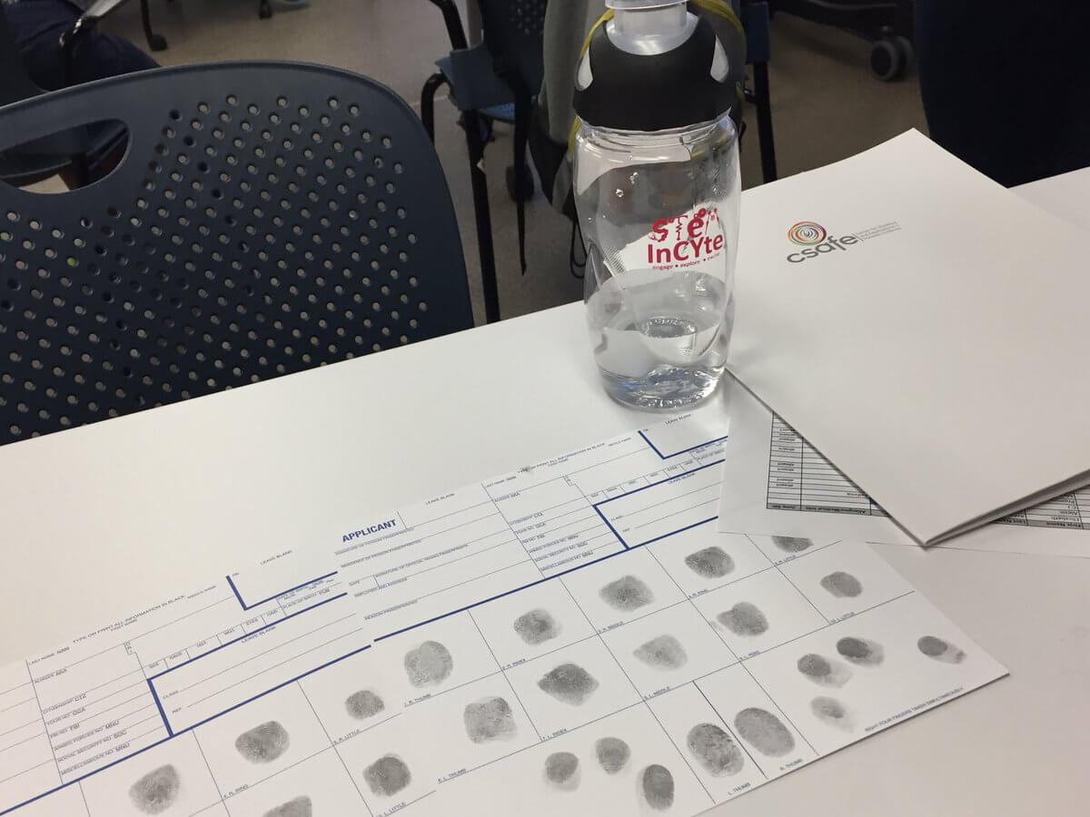 A water bottle and fingerprint forms on a table beside a white folder and a perforated chair.