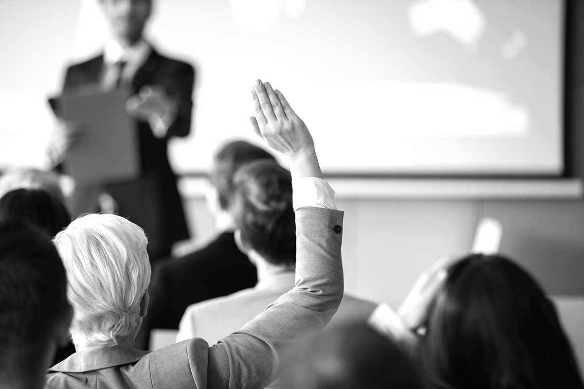 Person raising hand in an audience during a presentation; speaker in background holds a clipboard.